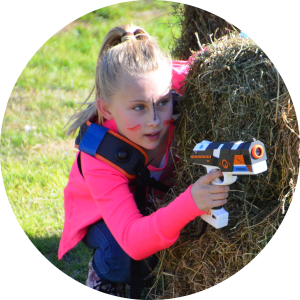 Girl with war paint and a pink sweater crouches behind a bale of hay. This is taken at an action birthday party for girls held at a farm in Dural